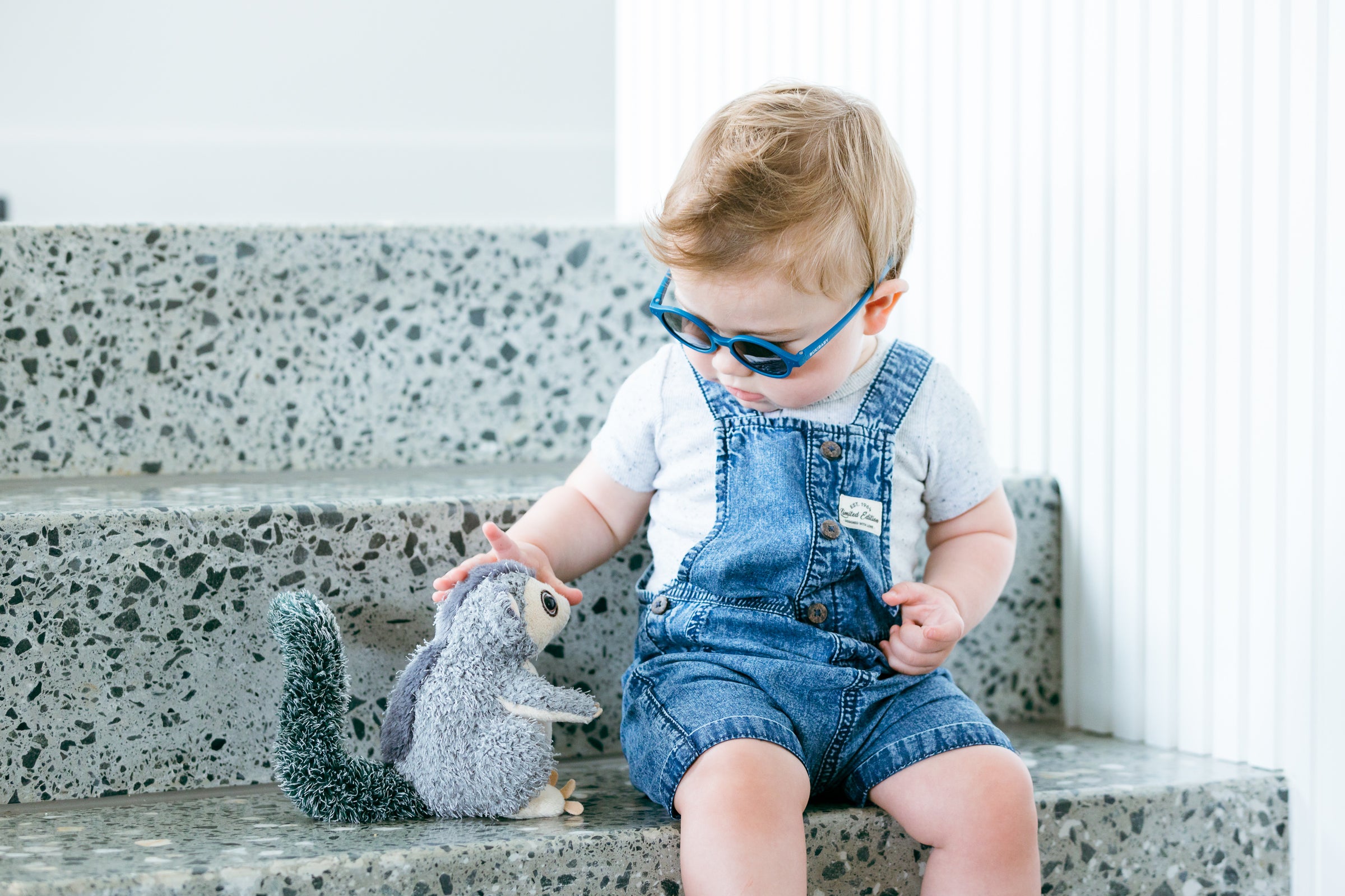 A curious toddler playing with their stuffed toy while wearing Bukibaby Sunglasses.