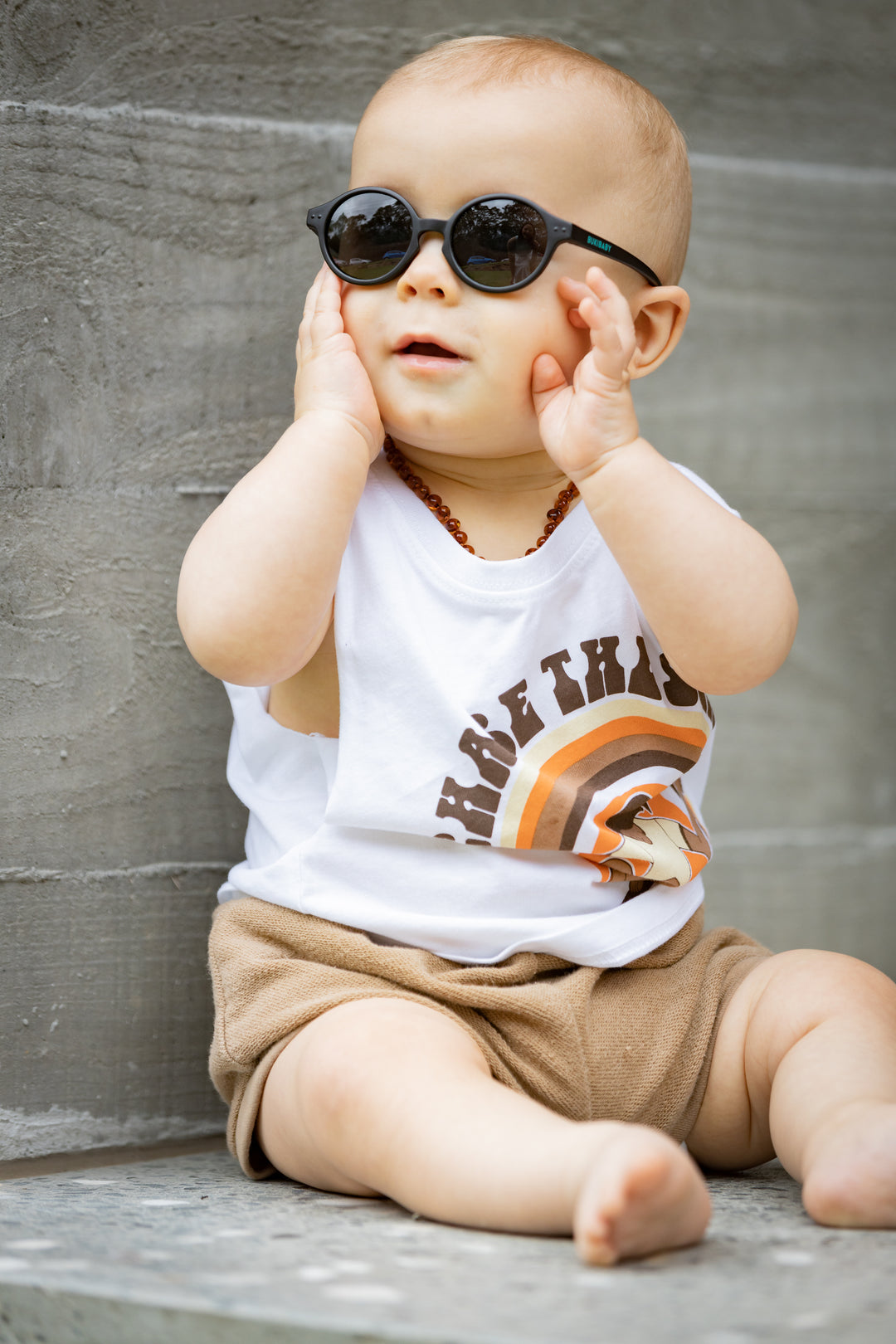 Toddler sitting on a outdoor sofa playing with his black Bukibaby Sunglasses.