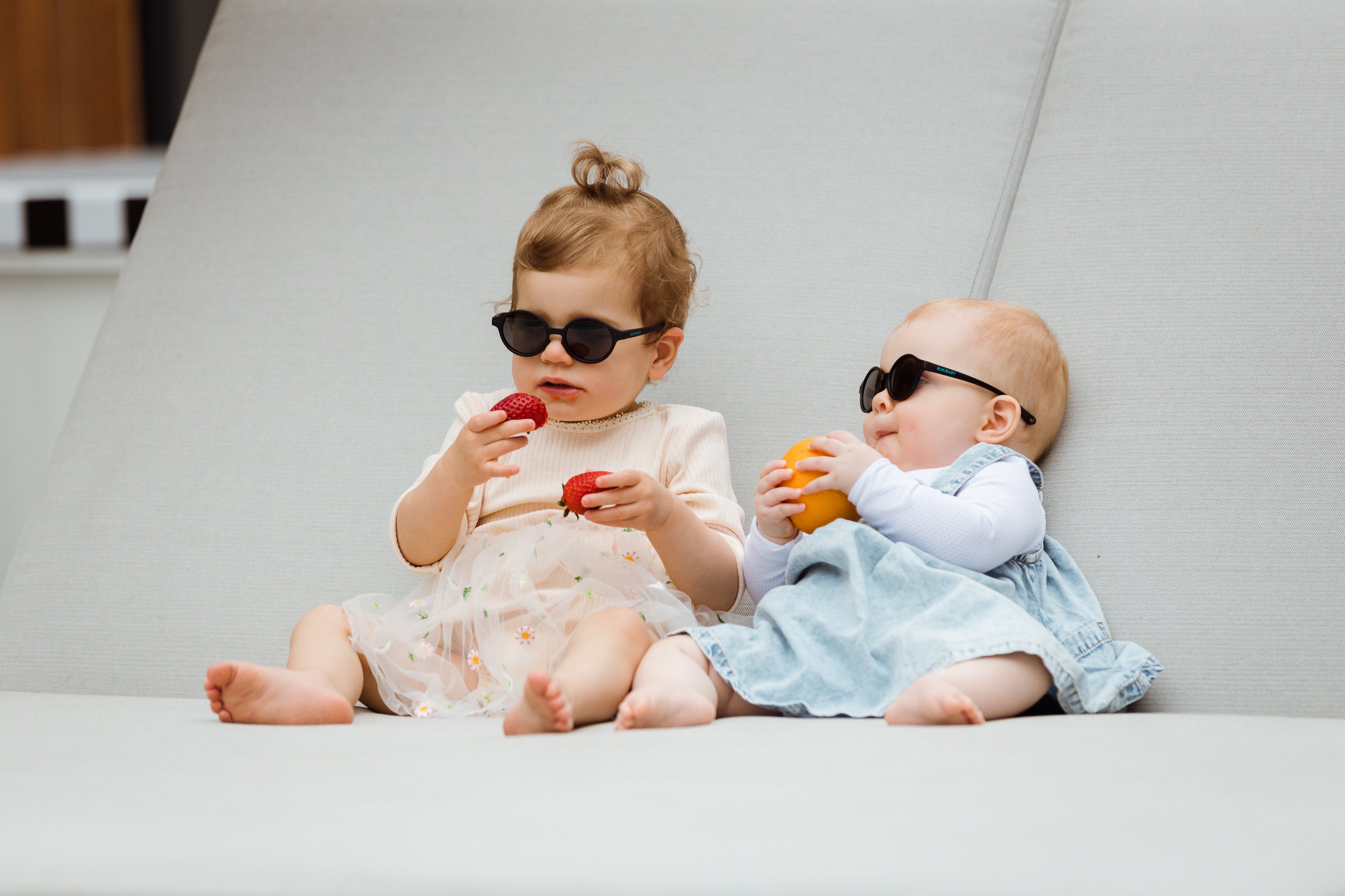 Two adorable babies eating fruit while sitting on an outdoor sofa and wearing Bukibaby Sunglasses