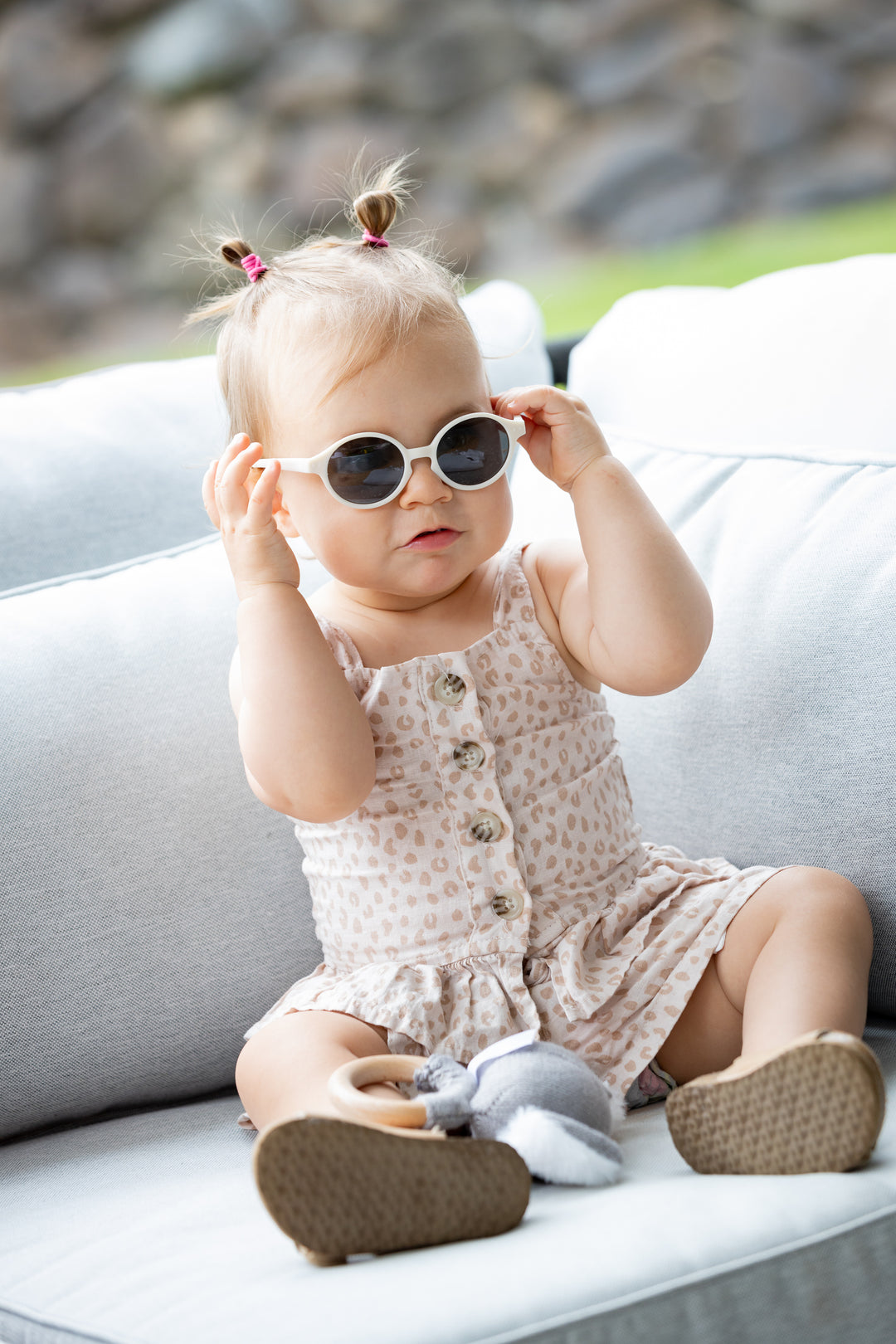 Toddler sitting on a outdoor sofa playing with her white Bukibaby Sunglasses.