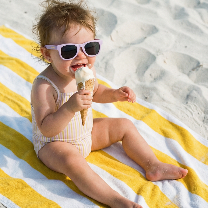 Child Baby wearing purple sunglasses at the beach, eating an ice cream cone.