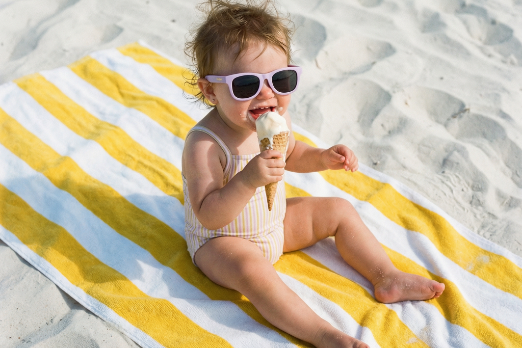 Child Baby wearing purple sunglasses at the beach, eating an ice cream cone.